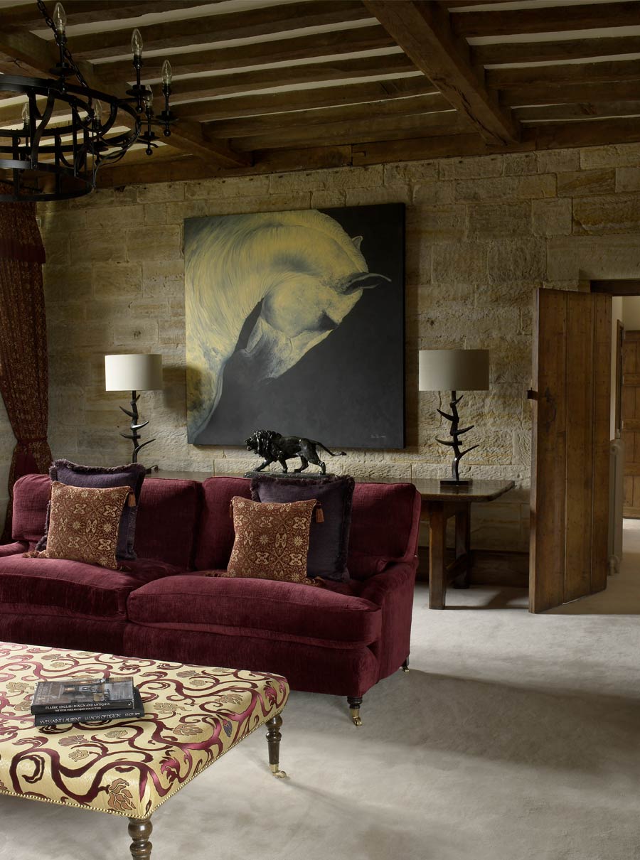 Plum velvet sofa in country house living room with statement table lamps and artwork against original stone walls.
