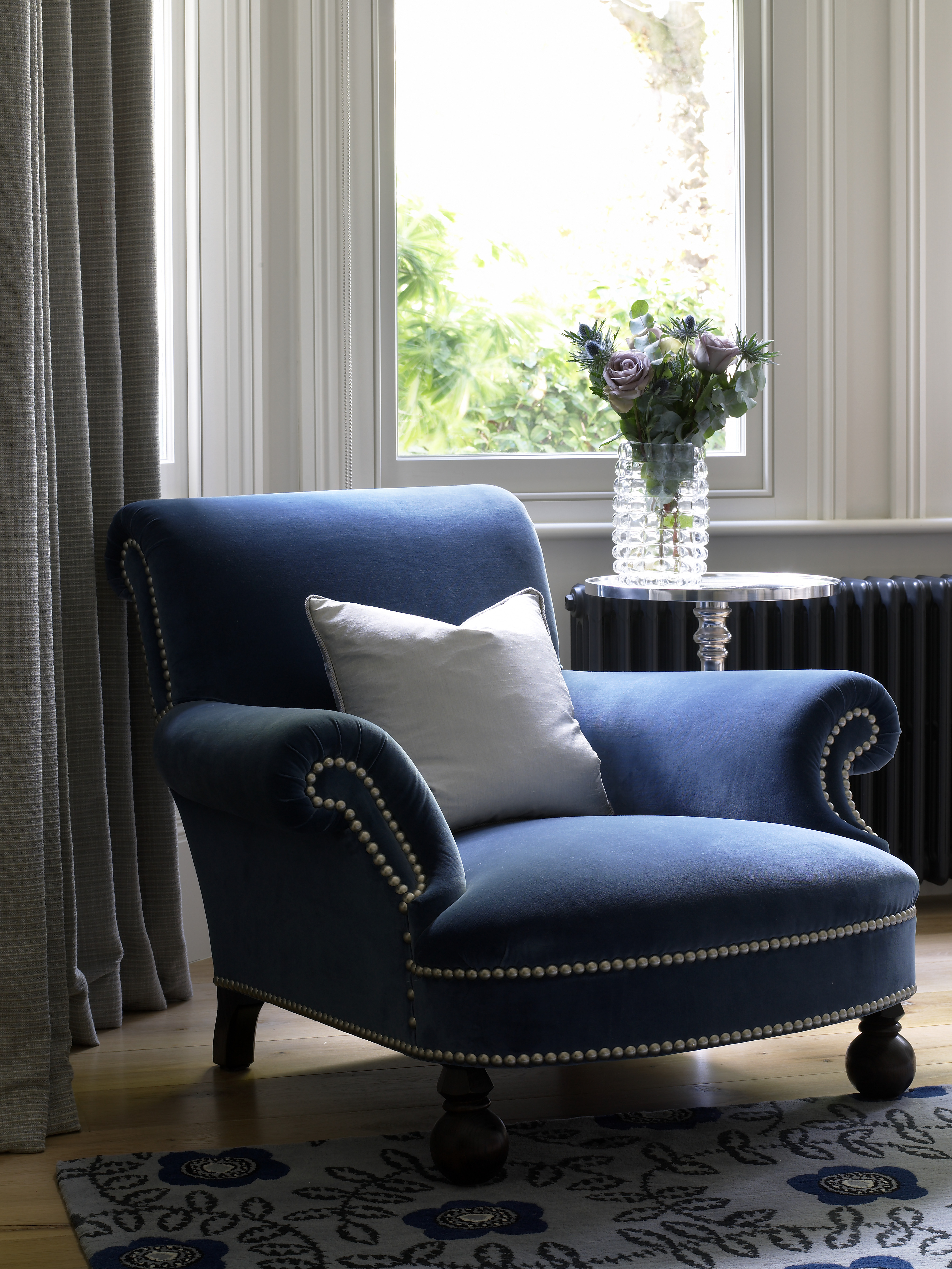 Midnight blue velvet armchair with scroll back and chrome studs beside contemporary patterned wool rug.